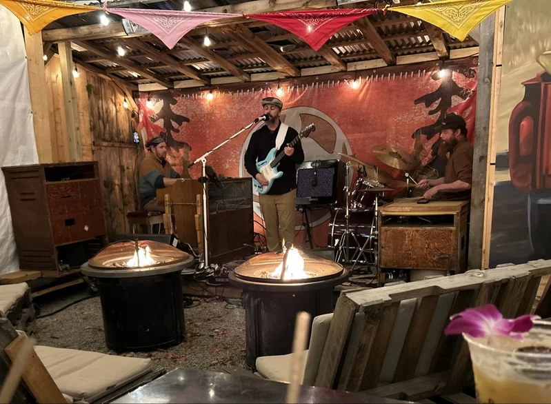 A live band on stage at Levitate Backyard in Marshfield, MA, with fire pits in front of the stage and string lights overhead